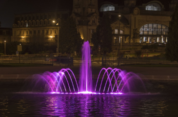 A purple fountain in the middle of a large lake or pond located near a school, university, or business.